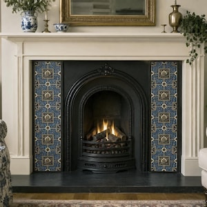 Puede incluir: Un inserto de chimenea de hierro fundido negro enmarcado por dos paneles verticales de azulejos azules decorativos con un patrón floral repetitivo en oro y blanco. La chimenea está situada dentro de una repisa blanca con fuego.