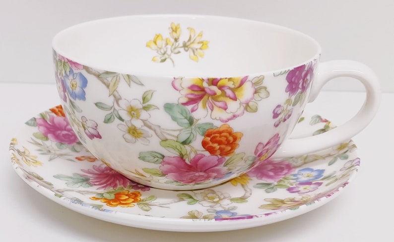 May include: A white teacup and saucer set with a floral pattern in shades of pink, orange, yellow, and blue. The floral design features a variety of flowers and leaves.