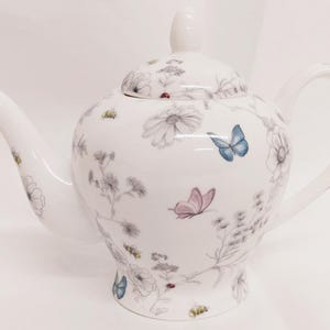 Pode incluir: Bule de porcelana branca com um design floral e de borboleta. A chaleira tem uma tampa e um bico.
