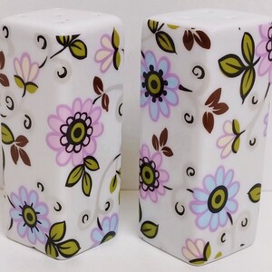 Könnte beinhalten: Ein Set aus zwei weißen Salz- und Pfefferstreuern aus Keramik mit floralem Design. Die Streuer haben einen weißen Hintergrund mit rosa, blauen und braunen Blumen und Blättern.