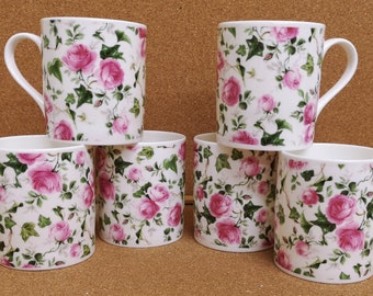 May include: Six white ceramic mugs with a pink rose floral pattern. The mugs have a handle on the right side.