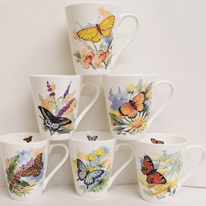 May include: Set of six white ceramic mugs with butterfly designs. Each mug features a different butterfly species with watercolor-like floral details. The mugs have a tall, tapered shape and a handle.