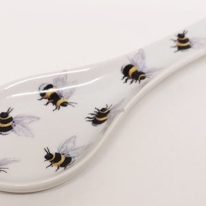 May include: White ceramic spoon rest with a bee design. The spoon rest is shaped like a spoon and has a handle. The bees are black and yellow and are scattered across the surface of the spoon rest.