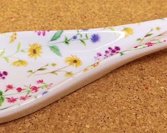 Könnte beinhalten: Weißer Keramik-Löffelhalter mit einem Blumenmuster mit gelben, rosa, violetten und grünen Blumen. Der Löffelhalter hat einen gebogenen Griff.