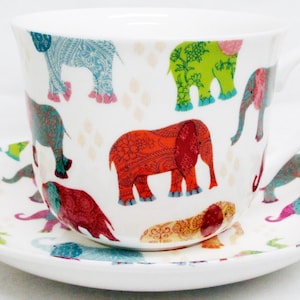 May include: A white ceramic teacup and saucer set. The cup and saucer are decorated with colourful elephant illustrations in shades of red, blue, green, and pink. The cup has a handle and the saucer is round.