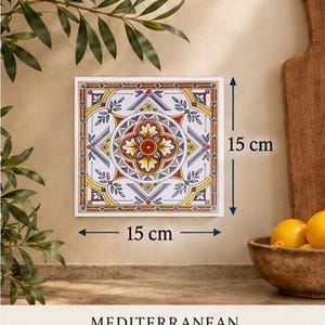 May include: A square decorative tile measuring 15 cm by 15 cm, featuring a symmetrical floral pattern in yellow, red, blue, and white. The design includes a central circular motif surrounded by geometric borders and leaf accents.