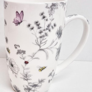 May include: White ceramic mug with a wraparound floral design in grey and black, featuring a pink butterfly, bees, and a ladybug. The mug has a large handle and is suitable for hot or cold beverages.