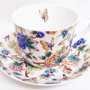 May include: White teacup and saucer set with a floral and butterfly design. The cup and saucer feature a colorful pattern of flowers, butterflies, and leaves in shades of blue, pink, orange, and yellow. The cup has a white handle.