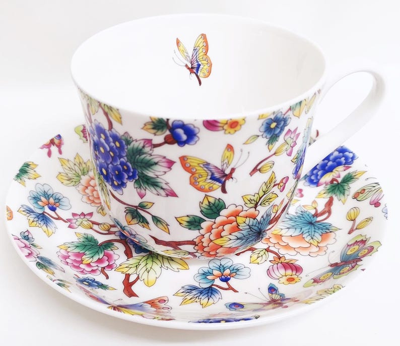 May include: White teacup and saucer set with a floral and butterfly design. The cup and saucer feature a colorful pattern of flowers, butterflies, and leaves in shades of blue, pink, orange, and yellow. The cup has a white handle.