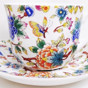 May include: White teacup and saucer set with a colorful floral and butterfly design. The cup and saucer feature blue, orange, yellow, and pink flowers and butterflies. The teacup has a curved handle.