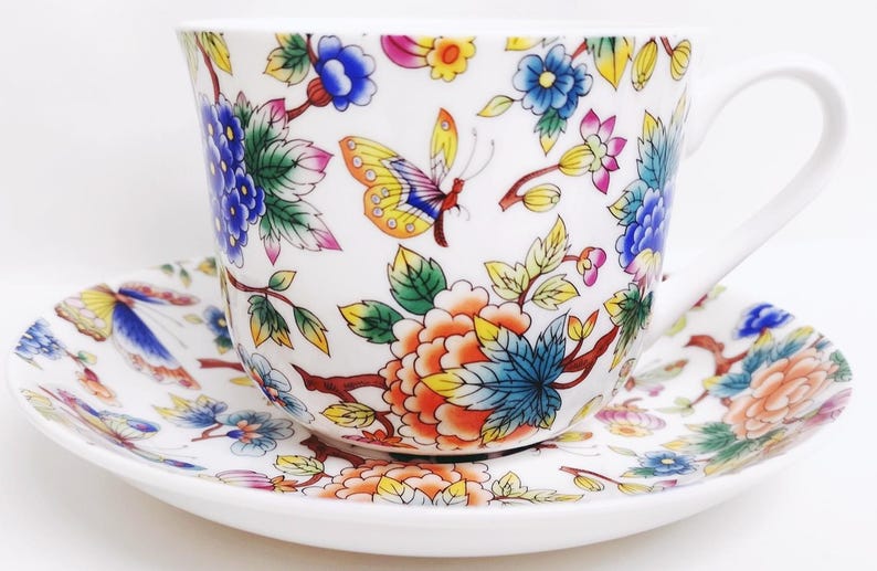 May include: White teacup and saucer set with a colorful floral and butterfly design. The cup and saucer feature blue, orange, yellow, and pink flowers and butterflies. The teacup has a curved handle.
