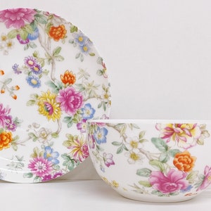 May include: A white teacup and saucer set with a floral pattern. The pattern features pink, yellow, orange, and blue flowers on a white background.