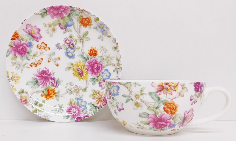 May include: A white teacup and saucer set with a floral pattern. The pattern features pink, yellow, orange, and blue flowers on a white background.
