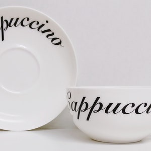 Peut inclure: Ensemble tasse et soucoupe en céramique blanche avec l'inscription "Cappuccino" en écriture cursive noire.