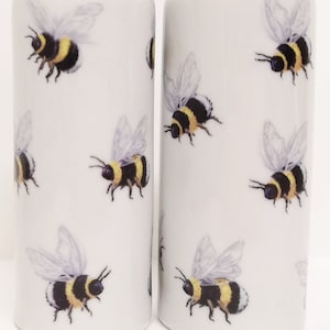 Può includere: Due saliere e pepiere in ceramica bianca. Ogni saliera e pepiera cilindrica è decorata con un motivo ripetuto di bombi. Le api sono nere e gialle con ali traslucide. Le saliere e le pepiere sono su sfondo bianco.