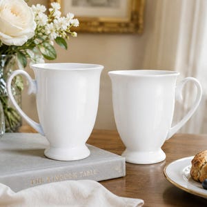 May include: Two matching white ceramic mugs with a smooth, glossy finish, featuring a flared rim, a curved handle, and a pedestal base. The mugs are plain, solid white, and have a classic, rounded silhouette designed for holding hot beverages.