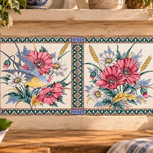 Könnte beinhalten: Zwei quadratische Keramikfliesen mit einem bemalten Motiv aus rosa Mohnblumen, weißen Gänseblümchen und Weizenähren. Die linke Fliese zeigt einen blau-gelben Vogel. Beide Fliesen sind von einer dekorativen Bordüre mit geometrischen Mustern umrahmt.