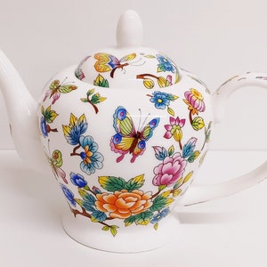 Peut inclure: Théière en porcelaine blanche avec un motif floral et papillon. La théière a un couvercle et une poignée. Les fleurs sont dans des tons de rose, orange, jaune, bleu et vert. Les papillons sont jaunes, verts et bleus.
