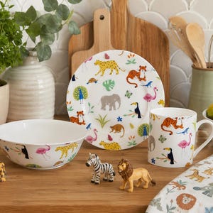 Può includere: Un set di stoviglie in ceramica bianca composto da un piatto, una ciotola e una tazza, decorati con illustrazioni colorate di vari animali selvatici tra cui una tigre, un elefante, un fenicottero, un tucano e un leopardo, con statuine di animali.