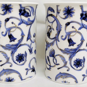 May include: Two white ceramic mugs with blue and gold floral designs. The mugs have a classic shape and a comfortable handle.