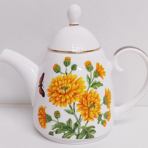 May include: White porcelain teapot with a floral design featuring yellow chrysanthemums and a butterfly. The teapot has a lid with a gold trim and a green handle.