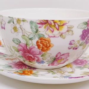 May include: A white teacup and saucer set with a floral pattern. The pattern features pink, orange, yellow, and purple flowers with green leaves on a white background.