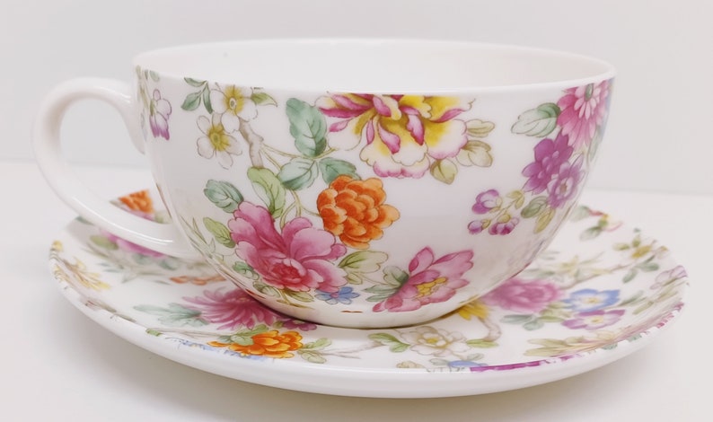 May include: A white teacup and saucer set with a floral pattern. The pattern features pink, orange, yellow, and purple flowers with green leaves on a white background.