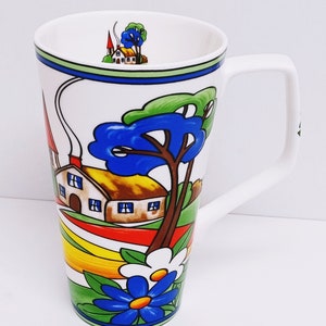 以下が含まれることがあります： カラフルなファンタジーな村のシーンが描かれた、背の高い白いセラミックマグカップ。デザインには、家、木、花、曲がりくねった道が描かれています。マグカップには大きな取っ手があります。