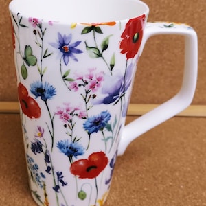 Op de afbeelding: Een hoge witte keramische mok met een bloemenpatroon. De mok heeft een verscheidenheid aan bloemen in tinten rood, blauw, roze en paars. De handgreep is wit en de mok is perfect om van een warme drank te genieten.