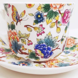 May include: White teacup and saucer set with a colorful floral and butterfly design. The cup and saucer are decorated with orange, pink, blue, and yellow flowers and green leaves. The teacup has a curved handle.