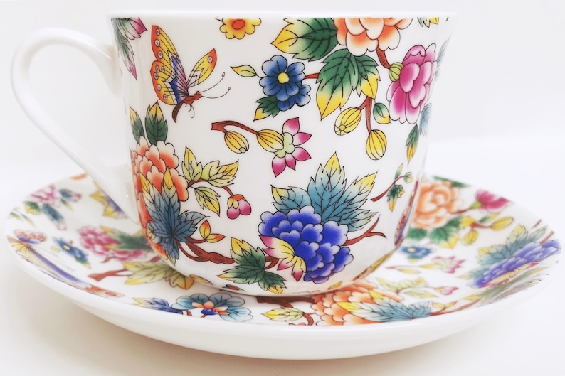 May include: White teacup and saucer set with a colorful floral and butterfly design. The cup and saucer are decorated with orange, pink, blue, and yellow flowers and green leaves. The teacup has a curved handle.