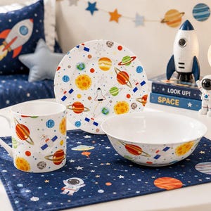 Peut inclure: Un mug, une petite assiette et un bol en céramique blanche, tous ornés d'un motif spatial coloré avec des planètes, des étoiles et des fusées. L'ensemble est disposé sur un set de table rectangulaire bleu foncé avec des étoiles et astronautes.