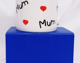 May include: White ceramic mug with a blue box. The mug has the word "Mum" written on it three times and three red hearts.