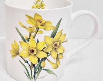May include: White ceramic mug featuring a vibrant illustration of yellow daffodils with green leaves. The mug has a curved handle and a floral design on the inside rim. The artist's signature, D. Hague, is visible on the mug.