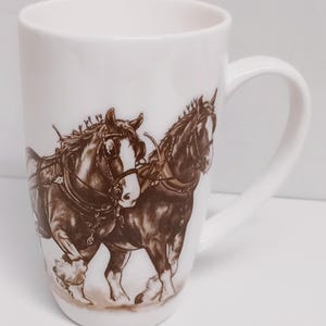 May include: White ceramic mug featuring a brown illustration of two horses pulling a cart. The horses are depicted walking with their heads turned towards the viewer.