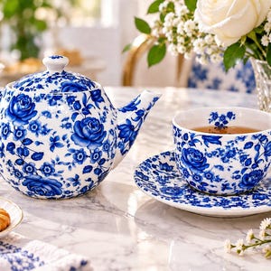 Könnte beinhalten: Ein Teekannen-, Tassen- und Untertassenset aus weißem Porzellan mit einem leuchtend blauen Blumenmuster aus Rosen und kleinen Blüten. Die Teekanne hat einen passenden Deckel, und die Tasse steht auf der Untertasse, beide mit dem gleichen blauen Design.