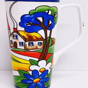 以下が含まれることがあります： 白いセラミック製のマグカップで、カラフルな家のイラスト、木々、花が描かれています。家は茶色の屋根と白い壁に青い窓が付いています。木は緑の葉と青い幹をしています。花は青と白で、中心は黄色です。