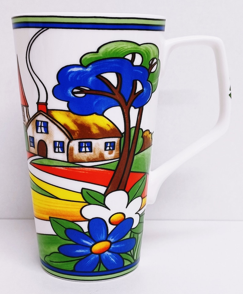 以下が含まれることがあります： 白いセラミック製のマグカップで、カラフルな家のイラスト、木々、花が描かれています。家は茶色の屋根と白い壁に青い窓が付いています。木は緑の葉と青い幹をしています。花は青と白で、中心は黄色です。
