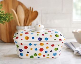 May include: A white ceramic butter dish with a lid, featuring a hand-painted pattern of multicolored polka dots in shades of red, blue, green, yellow, and purple. The rectangular container has rounded corners and a small knob handle on the lid.