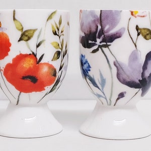 May include: Two white ceramic egg cups with floral designs. One cup features red poppies and a bee, the other has purple and blue flowers. Both have a pedestal base.