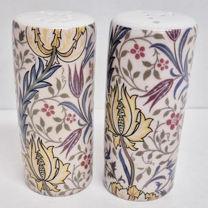 Op de afbeelding: Een paar keramische zout- en peperstrooiers met een bloemmotief. De strooiers zijn cilindrisch met een witte bovenkant en een crèmekleurige body. De body is versierd met gele bloemen, blauwe bladeren en rode bessen.