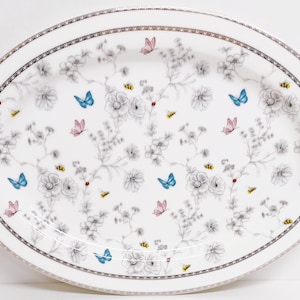 Könnte beinhalten: Weiße ovale Servierplatte mit silbernem Rand und einem Blumenmuster mit grauen Blumen, blauen und rosa Schmetterlingen und gelben Bienen.