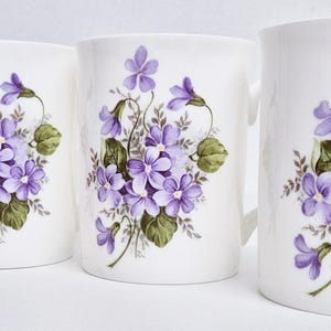 May include: Four white ceramic mugs with a floral design. Each mug features a cluster of purple violets with green leaves. The mugs are cylindrical with a handle on one of the mugs.