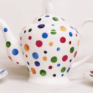 Può includere: Una teiera bianca e due tazze da tè con piattini abbinati. La teiera e le tazze sono decorate con un motivo a pois colorati in tonalità di blu, verde, rosso, giallo e arancione.