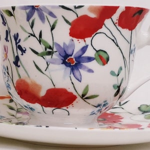 Op de afbeelding: Een wit theekopje en schotelset met een bloemenpatroon met rode klaprozen, blauwe bloemen en groene bladeren.