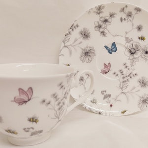 May include: A white teacup and saucer set with a floral pattern featuring butterflies and bees. The cup and saucer are decorated with a delicate gray and white floral design, with pink and blue butterflies and yellow bees.