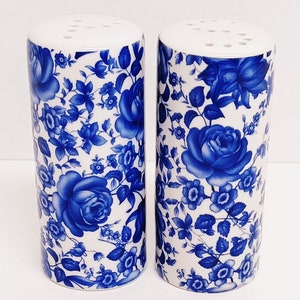 May include: A set of two white ceramic salt and pepper shakers with a blue floral design. The shakers have a cylindrical shape and a small hole on the top for dispensing salt and pepper.