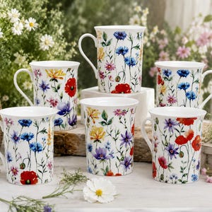 Peut inclure: Un ensemble de cinq tasses en céramique blanche, chacune ornée d'un imprimé botanique coloré de fleurs sauvages, notamment des coquelicots rouges, des bleuets bleus et des fleurs jaunes. Les tasses ont une forme cylindrique avec une anse.