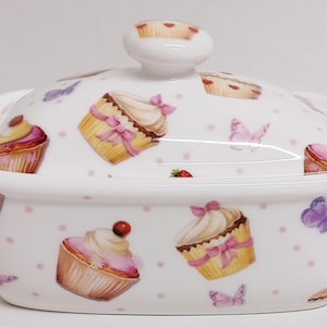 May include: White ceramic butter dish with a lid. The dish and lid are decorated with a pink and white polka dot pattern and images of cupcakes and butterflies.