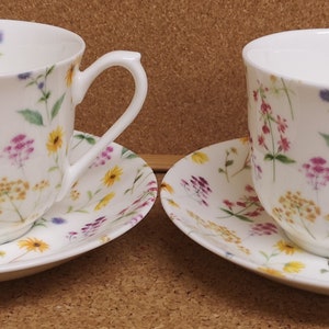 May include: Two white porcelain teacups and saucers featuring a floral design. The cups and saucers are adorned with a pattern of colorful wildflowers in yellow, pink, purple, and green. Each cup has a curved handle.